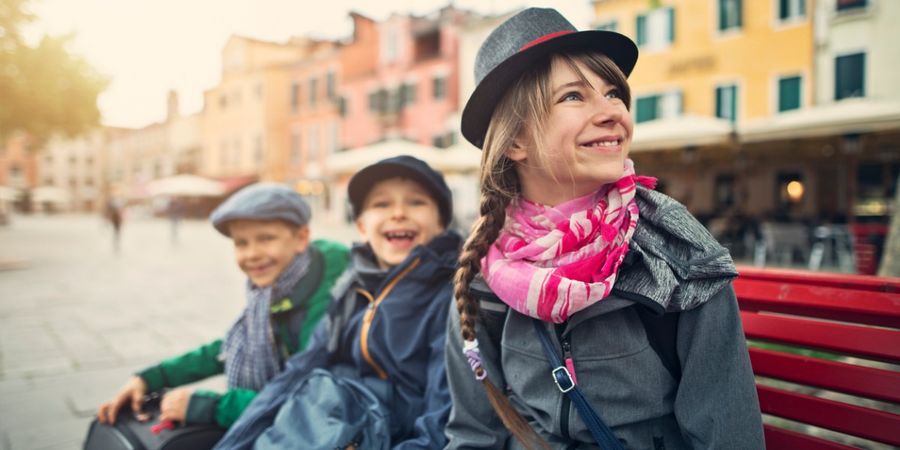 A photo of three kids sitting on a bench in front of brightly coloured buildings.