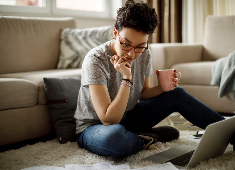 A woman sitting on the floor in front of her laptop, holding a pen in one hand and a coffee cup in the other.