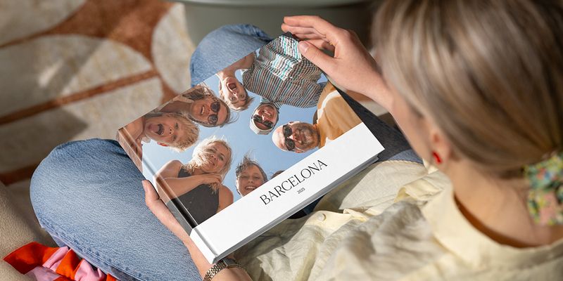 A woman looks at a photo book titled 'Barcelona', featuring an image of lots of smiling family faces looking down into the camera, illustrating Photobox's guide to making a group holiday photo book.